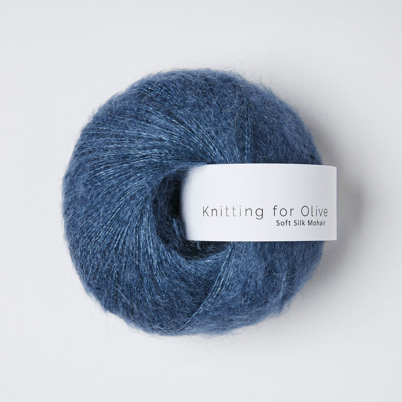 “Bol wol in de kleur ‘Blue Jeans’ van Knitting for Olive, uit de Soft Silk Mohair-lijn, luchtig en glanzend opgerold met een wit label tegen een witte achtergrond – luxe mohair-zijde garen in denimblauw.”