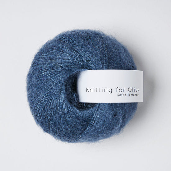 “Bol wol in de kleur ‘Blue Jeans’ van Knitting for Olive, uit de Soft Silk Mohair-lijn, luchtig en glanzend opgerold met een wit label tegen een witte achtergrond – luxe mohair-zijde garen in denimblauw.”