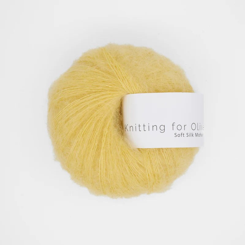 “Bol wol in de kleur ‘Buttercup’ van Knitting for Olive, uit de Soft Silk Mohair-lijn, pluizig en luchtig opgerold met een wit label tegen een witte achtergrond – luxe en duurzaam garen van mohair en zijde.”
