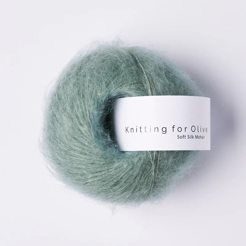 “Bol wol in de kleur ‘Dusty Aqua’ van Knitting for Olive, uit de Soft Silk Mohair-lijn, pluizig en luchtig opgerold met een wit label tegen een witte achtergrond – luxe en duurzaam garen van mohair en zijde.”