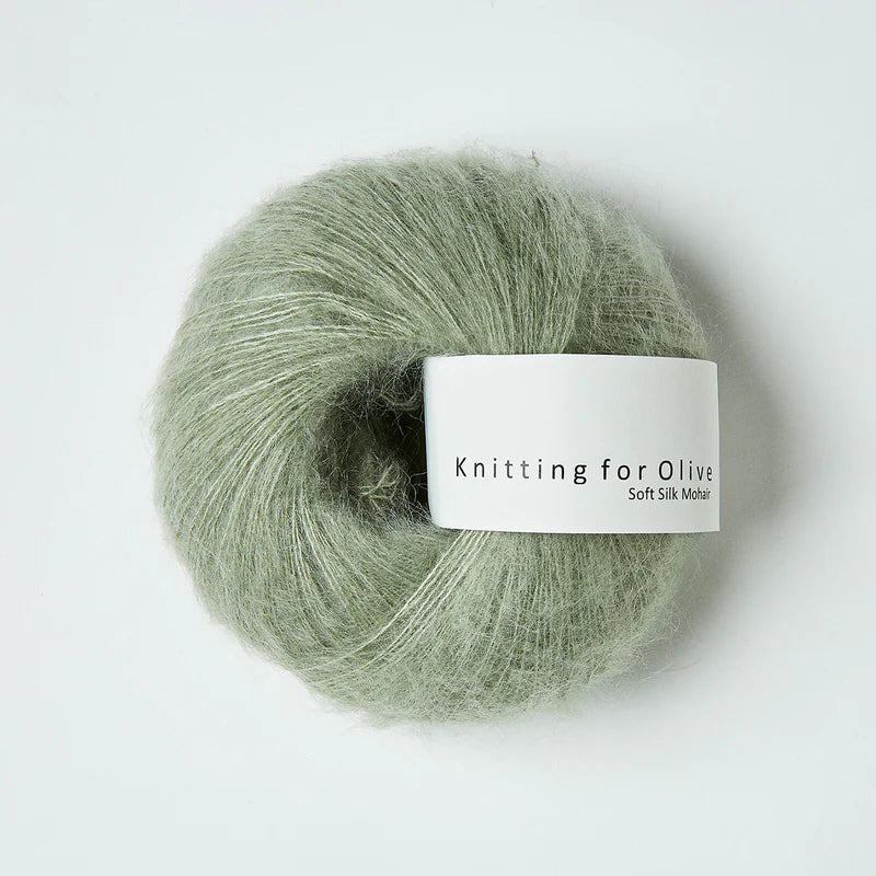 “Bol wol in de kleur ‘Dusty Artichoke’ van Knitting for Olive, uit de Soft Silk Mohair-lijn, pluizig en luchtig opgerold met een wit label tegen een witte achtergrond – luxe en duurzaam garen van mohair en zijde.”