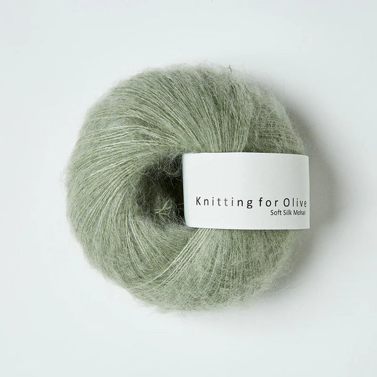 “Bol wol in de kleur ‘Dusty Artichoke’ van Knitting for Olive, uit de Soft Silk Mohair-lijn, pluizig en luchtig opgerold met een wit label tegen een witte achtergrond – luxe en duurzaam garen van mohair en zijde.”
