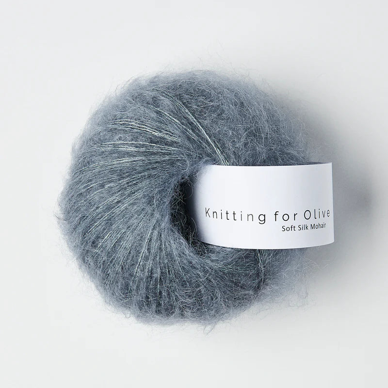“Bol wol in de kleur ‘Dusty Petrol’ van Knitting for Olive, uit de Soft Silk Mohair-lijn, pluizig en luchtig opgerold met een wit label tegen een witte achtergrond – luxe en duurzaam garen van mohair en zijde.”