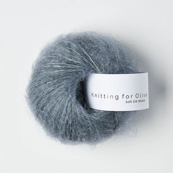 “Bol wol in de kleur ‘Dusty Petrol’ van Knitting for Olive, uit de Soft Silk Mohair-lijn, pluizig en luchtig opgerold met een wit label tegen een witte achtergrond – luxe en duurzaam garen van mohair en zijde.”