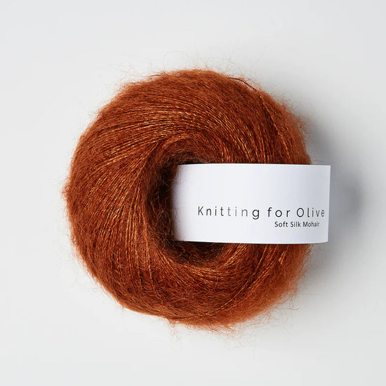 “Bol wol in de kleur ‘Rust’ van Knitting for Olive, uit de Soft Silk Mohair-lijn, pluizig en luchtig opgerold met een wit label tegen een witte achtergrond – luxe en duurzaam garen van mohair en zijde.”