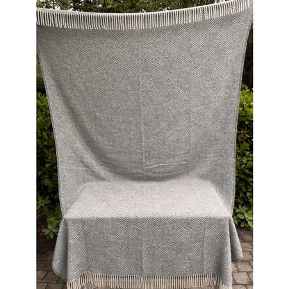 Groot wollen plaid in grijs-beige met patroon en franjes, volledig uitgehangen buiten tegen een achtergrond van groene struiken.