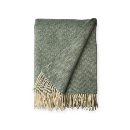 Groen wollen plaid met subtiel visgraatpatroon en lichte franjes, deels omgeslagen zodat de zachte structuur en afwerking zichtbaar zijn.