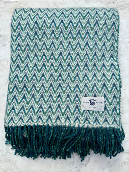 Opgevouwen blauw-wit wollen plaid met zigzagpatroon en franjes, liggend op een witte ondergrond. Het label van Belpo Textile is zichtbaar aan de rand.