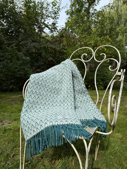 Blauw-wit wollen plaid met zigzagpatroon, gedrapeerd over een sierlijke witte tuinstoel in een groene tuinomgeving.