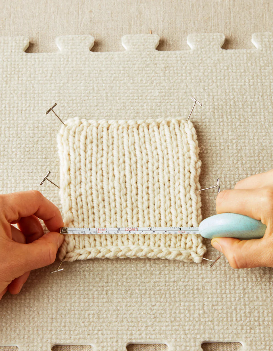 Handen die een gebreid lapje opspannen met Cocoknits T-pins op een blokmat tijdens het meten van steken.