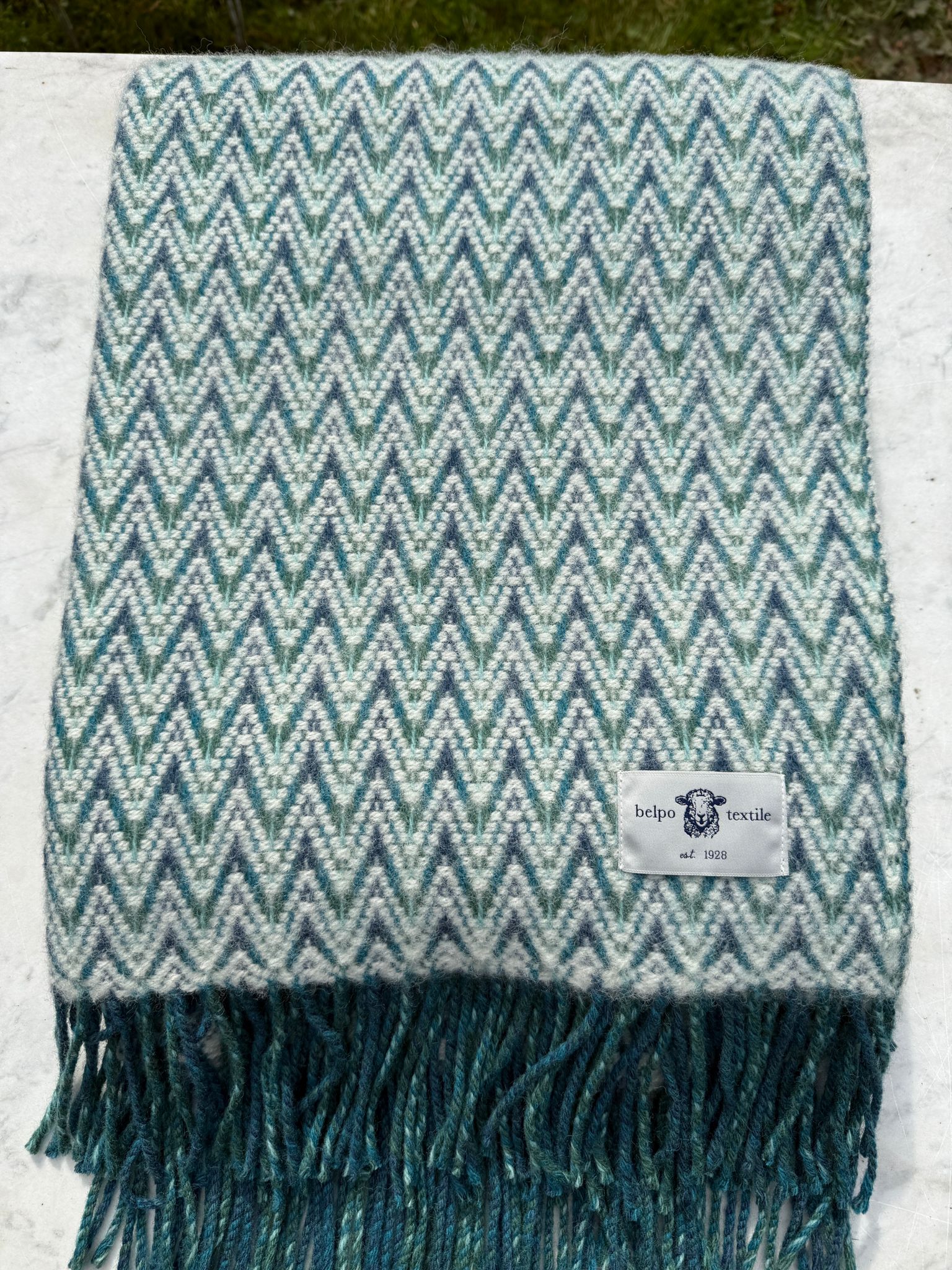 Gevouwen blauw-wit plaid van wol met zigzagweefsel en franjes, liggend op marmeren ondergrond. Het label van Belpo Textile is goed zichtbaar.