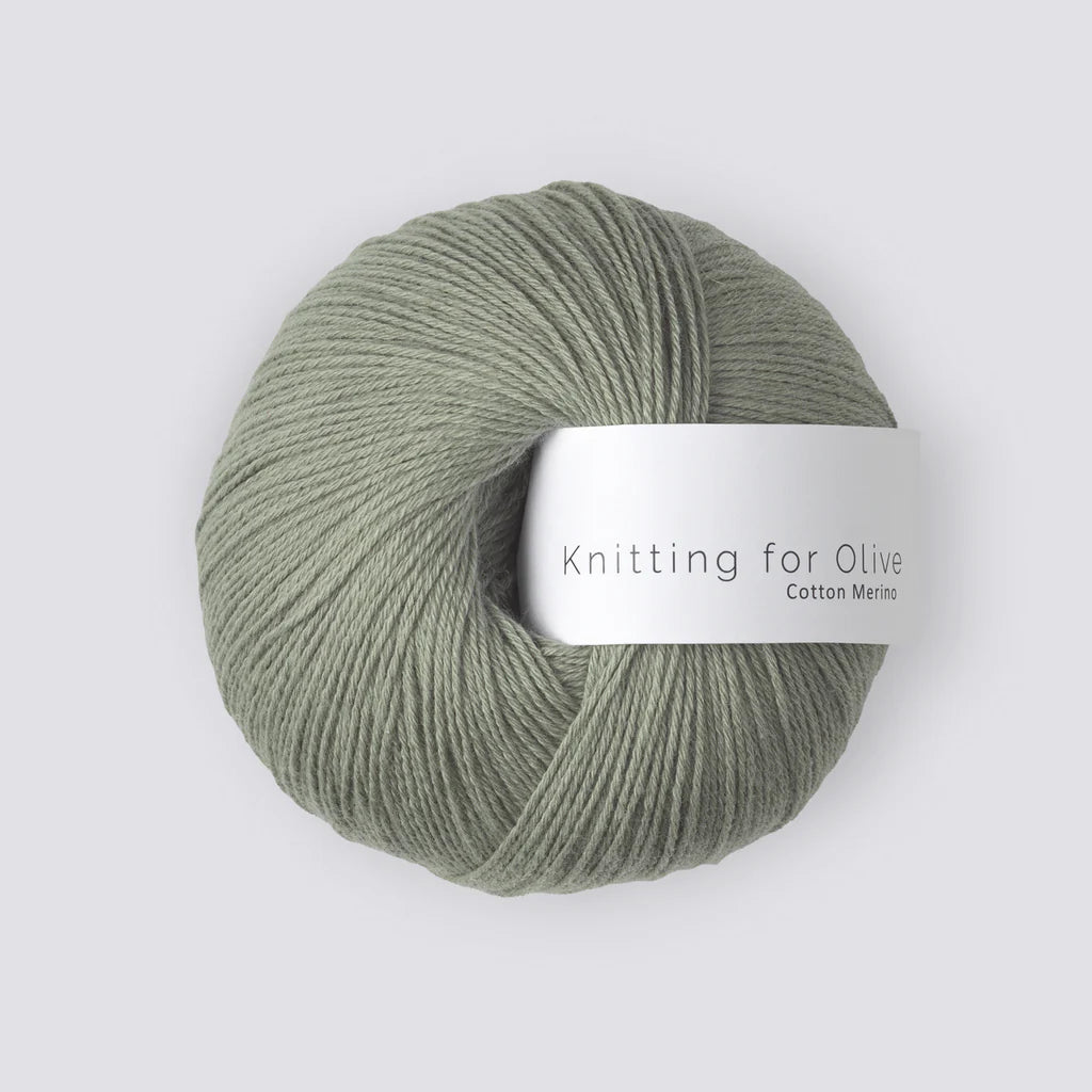 “Bol zachte wol in de kleur ‘Artichoke’ van het merk Knitting for Olive, uit de Cotton Merino-lijn, mooi opgerold met een wit label tegen een rustige, lichtgrijze achtergrond – perfect voor breiprojecten met een natuurlijke uitstraling.”