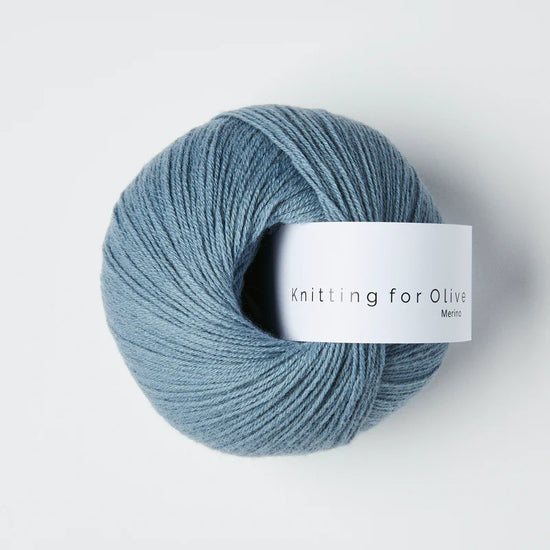 “Bol wol in de kleur ‘Dusty Dove Blue’ van Knitting for Olive, uit de Merino-lijn, netjes opgerold met wit label tegen een witte achtergrond – zacht, ademend en duurzaam garen.”