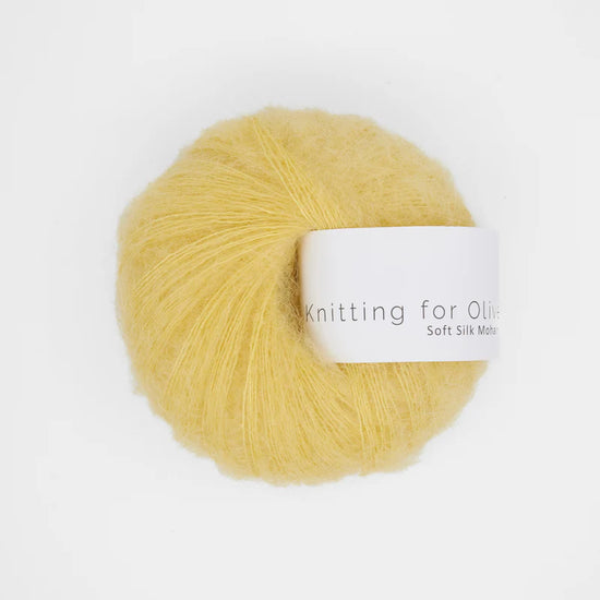 “Bol wol in de kleur ‘Buttercup’ van Knitting for Olive, uit de Soft Silk Mohair-lijn, pluizig en luchtig opgerold met een wit label tegen een witte achtergrond – luxe en duurzaam garen van mohair en zijde.”