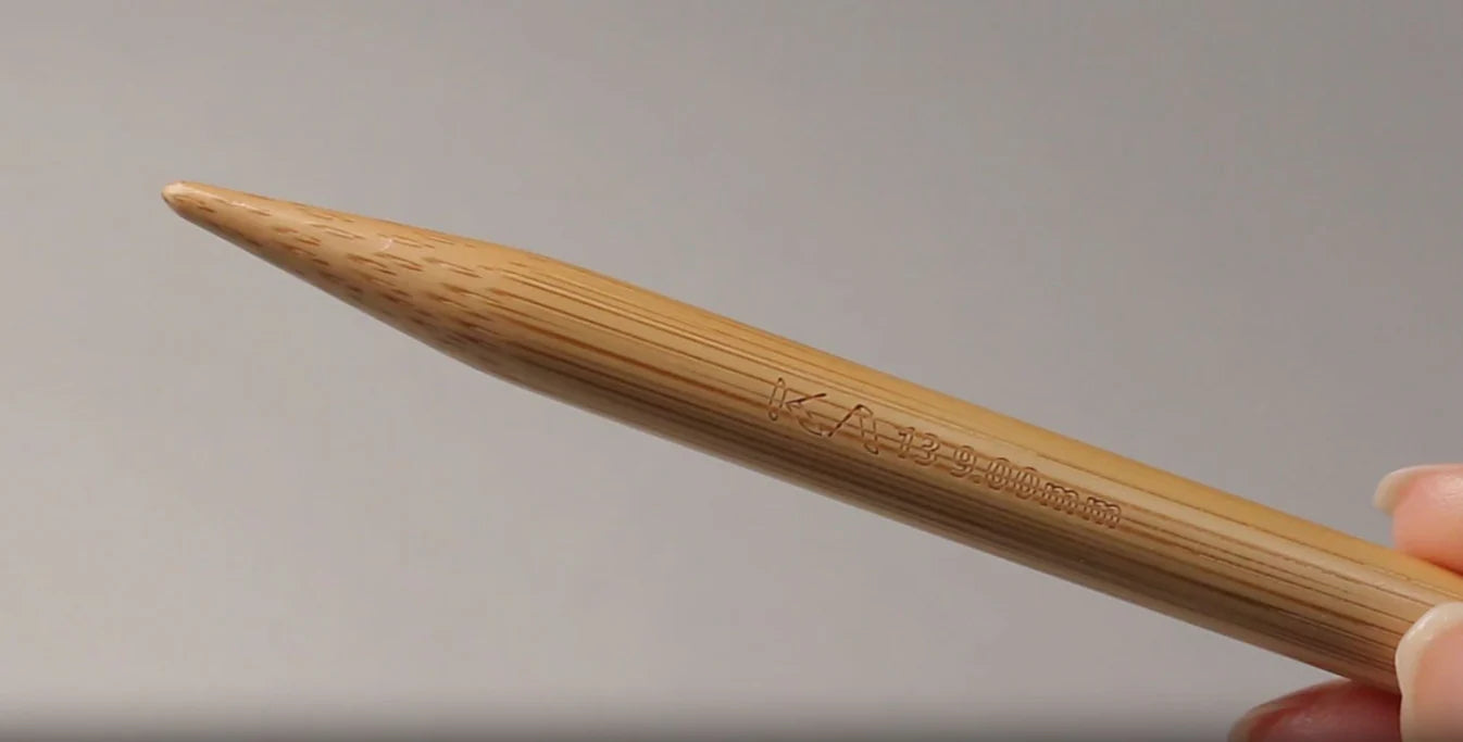 Close-up van een Seeknit bamboe breinaaldpunt met lasergegraveerde maat en logo. De gladde afwerking en natuurlijke bamboevezelstructuur zijn duidelijk zichtbaar, vastgehouden tussen duim en wijsvinger.