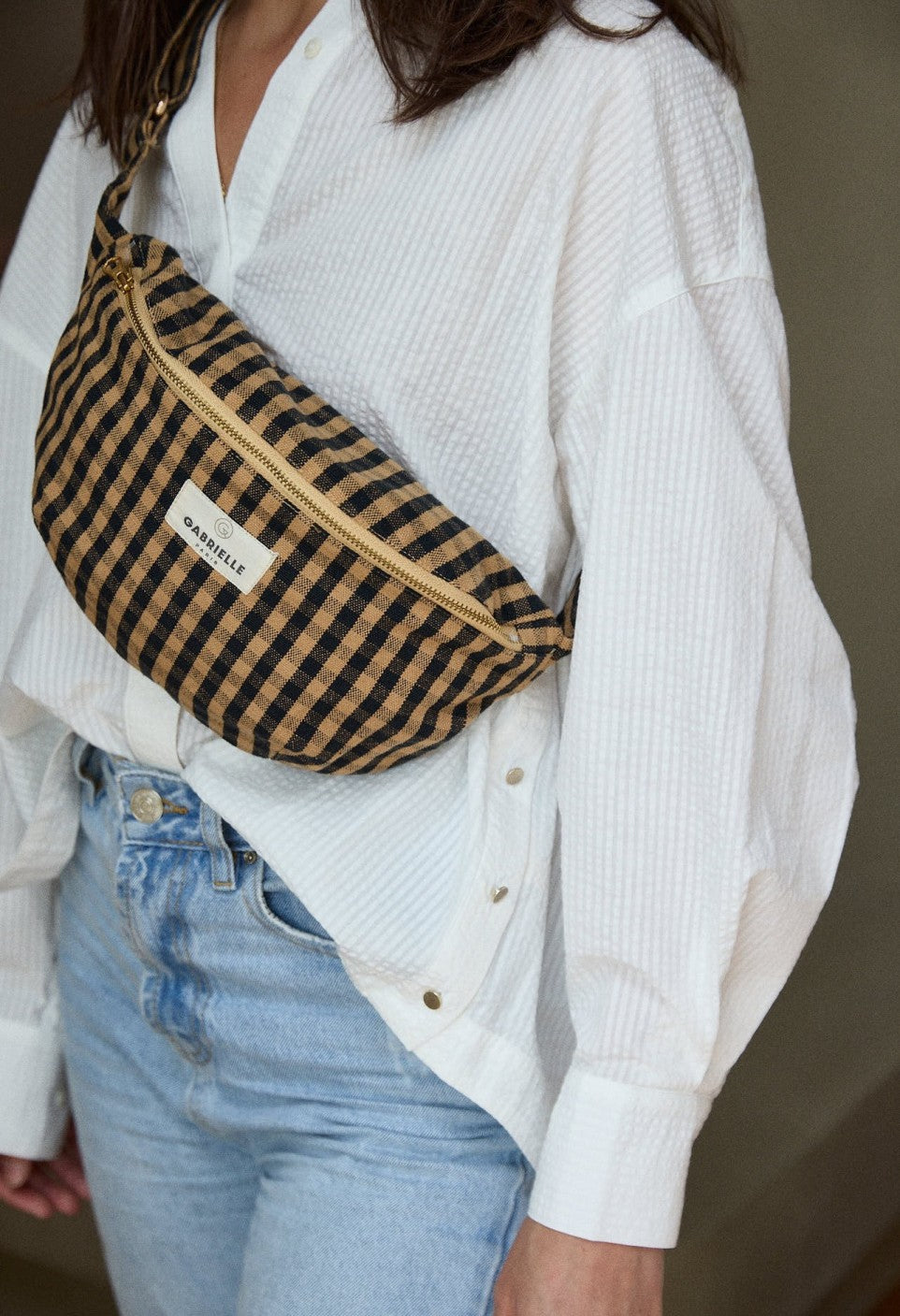 Close-up van Gabrielle Paris Waistbag in Vichy Café, gedragen over een witte blouse. Beige-bruin geruite heuptas van katoen met gouden rits en label.