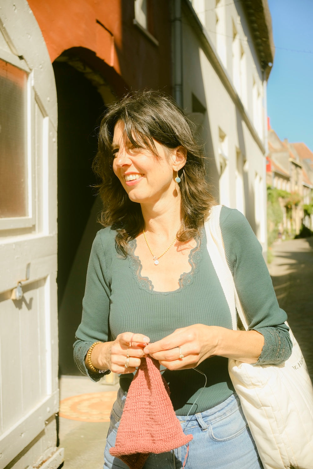 Christel van Ons Lykke, al wandelend aan het breien met oudroze garen in een zonnig straatje in Zutphen – een sfeerbeeld van ontspannen handwerk.